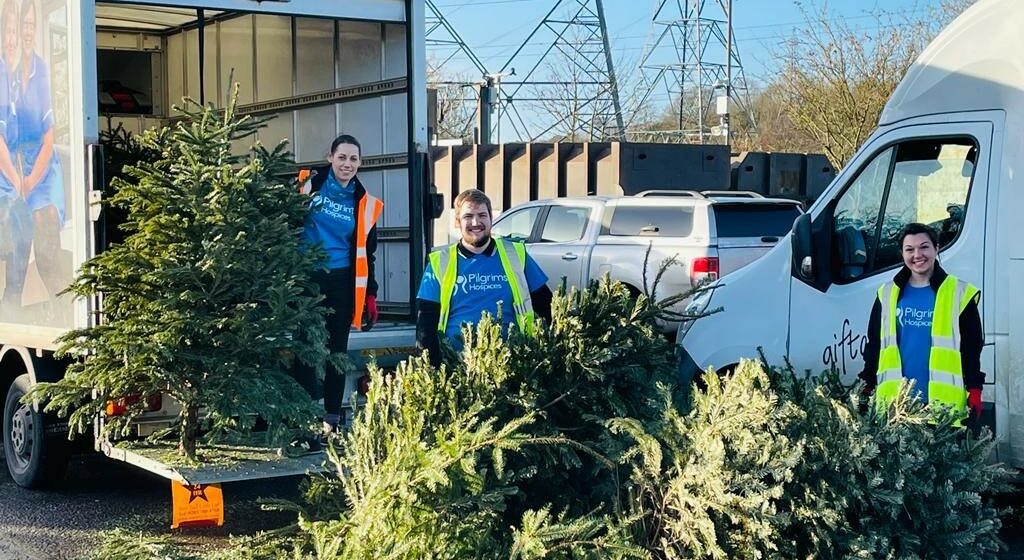 Tree Recycling Crews A group of three people wearing Hi Vis jackets and blue Pilgrims Hospices t-shirts, stood in front of vans and with real Christmas trees.