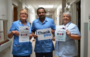 A group of three healthcare workers wearing blue uniforms, holding up paper signs; the middle sign reads: Thank you and the other two signs include text about Gift Aid.