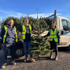 A group of three people wearing Hi Vis jackets, standing in front of a van and with real Christmas trees.